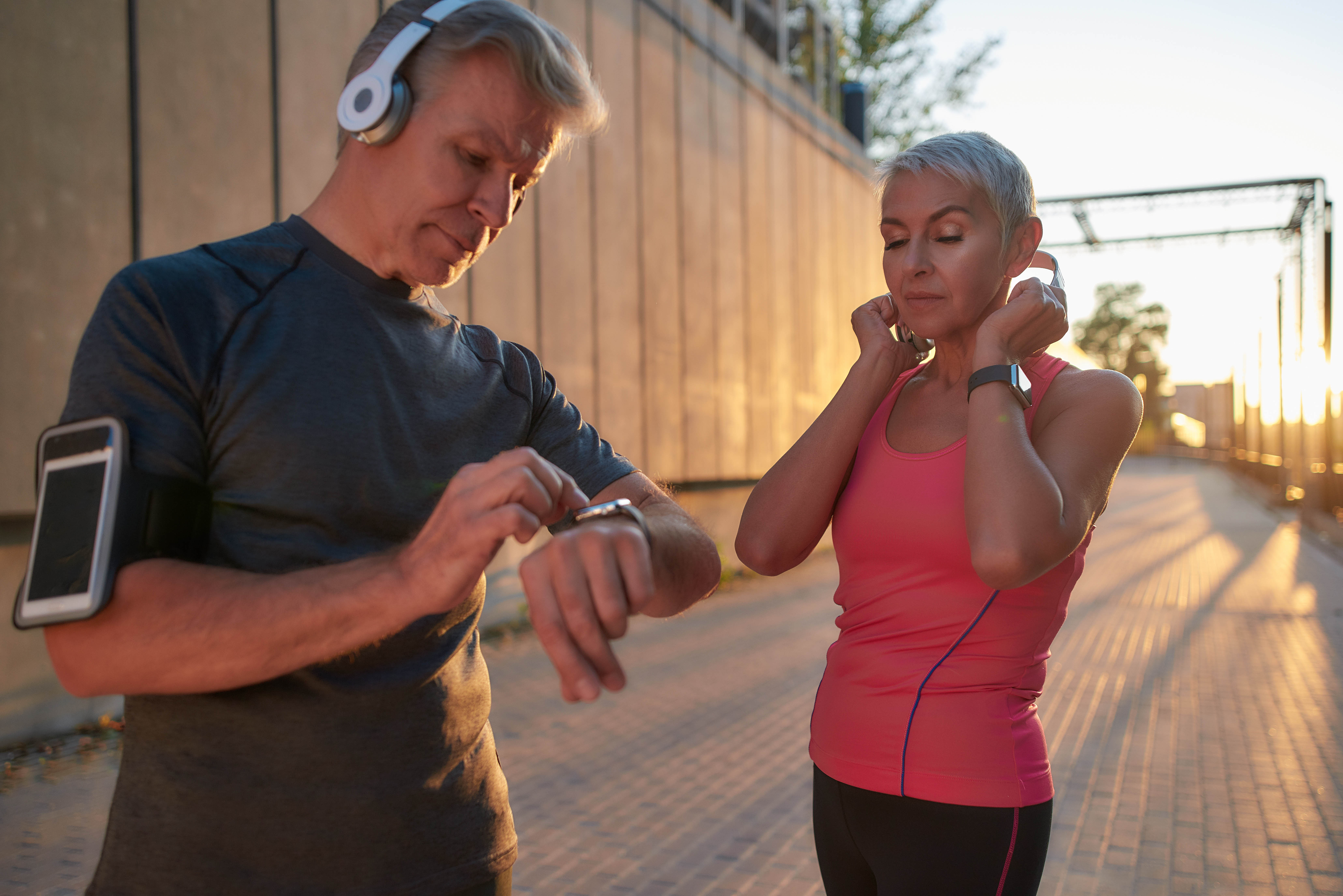 Older couple preparing for a run. He's wearing headphones and setting his smart watch. She has her eyes closed and is putting on her headphones, wearing a pink shirt.